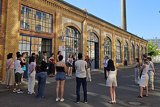Personen stehen im Kreis und schauen sich an. Im Hintergrund ein altes Industriegebäude mit Schornstein