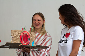Drei Frauen stehen nebeinander, die mittlere hält ein Modell einer Ausstellung mit rotem Eingansportal